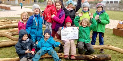 Eine Gruppe Kinder sitzt auf einem Baumstamm. Die Kinder halten einen Scheck des Deutschen Kinderhilfswerkes. 