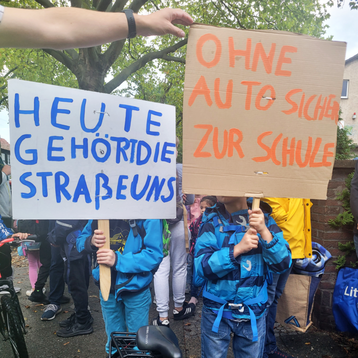 Zwei Kinder halten selbstgebastelte Schilder hoch. Auf denen ist zu lesen: Heute gehört die Straße uns. Und: Ohne Auto sicher zur Schule. 