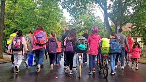 Eine Gruppe aus Kindern und Erwachsenen läuft gemeinsam eine Straße entlang. Einige von ihnen tragen Schulranzen oder schieben Fahrräder.