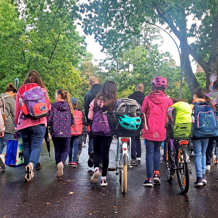 Eine Gruppe aus Kindern und Erwachsenen läuft gemeinsam eine Straße entlang. Einige von ihnen tragen Schulranzen oder schieben Fahrräder.