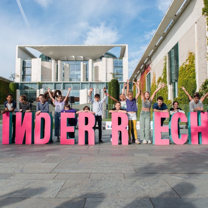 Pinke Buchstaben stehen vor dem Bundeskanzleramt in Berlin. Sie bilden das Wort "Kinderrechte". Dahinter stehen Kinder und strecken ihre Arme in die Luft. 