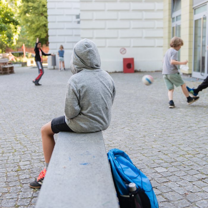 Ein Junge sitzt auf einer Mauer. Er trägt einen grauen Kapuzenpulli und hat die Kapuze über den Kopf gezogen. Vor ihm spielen Kinder Fußball.