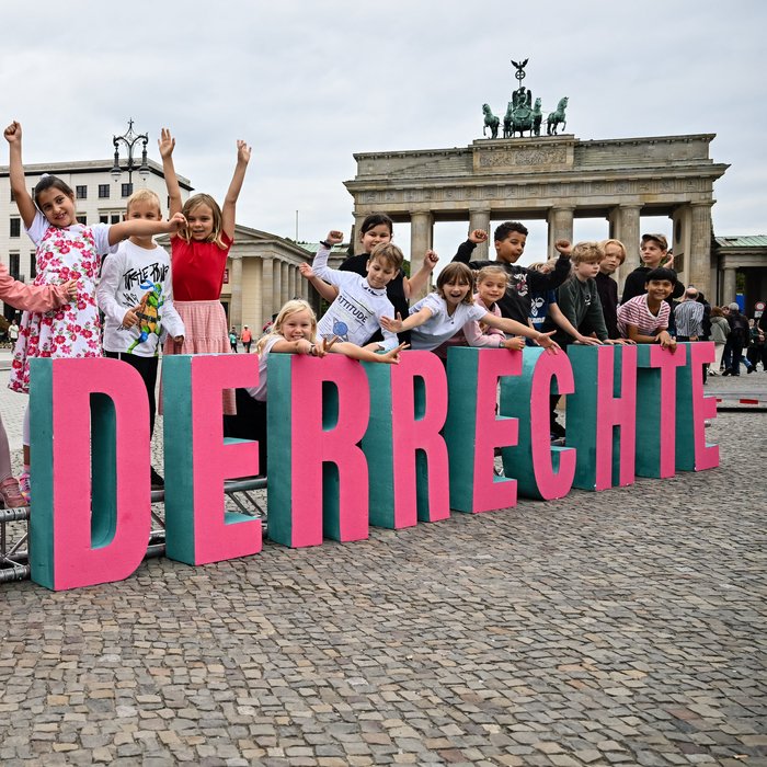Vor dem Brandenburger Tor bilden große Buchstaben das Wort "Kinderrechte". Dahinter stehen Kinder und jubeln. 