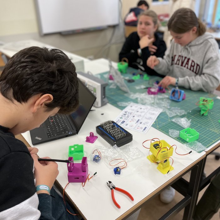 Drei Schüler*innen sitzen an einem Tisch und arbeiten mit Werkzeugen und einem Laptop.