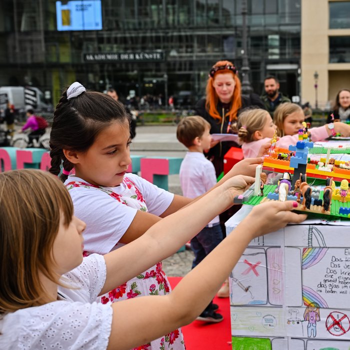 Kinder legen ihre bebauten Legoplatten auf ein aus Karton gebautes Haus.