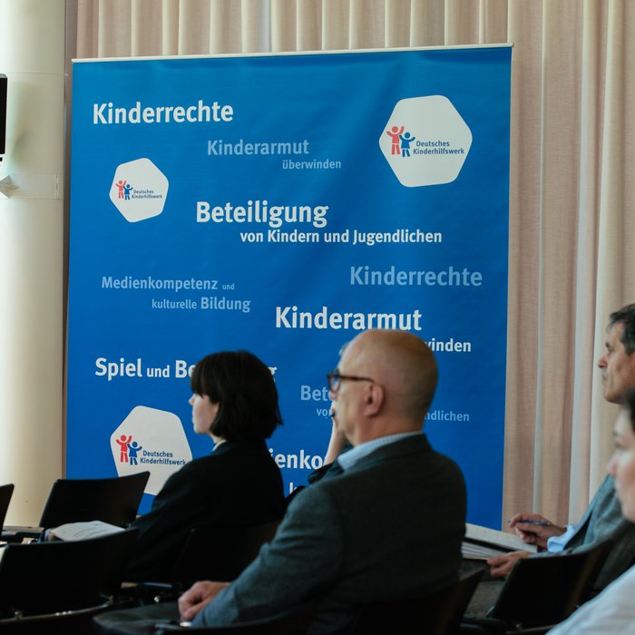 Mehre Menschen sitzen in einem Raum. Hinter ihnen ist die Pressewand des Deutschen Kinderhilfswerkes zu sehen. Darauf stehen Worte wie "Beteiligung" und "Kinderrechte".
