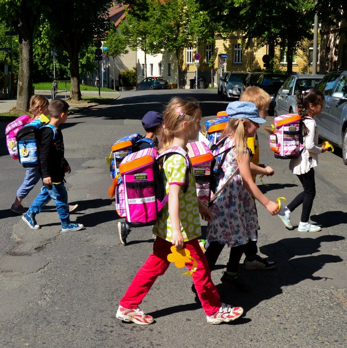Mehrere Kinder mit Schulranzen überqueren gemeinsam eine Straße. 