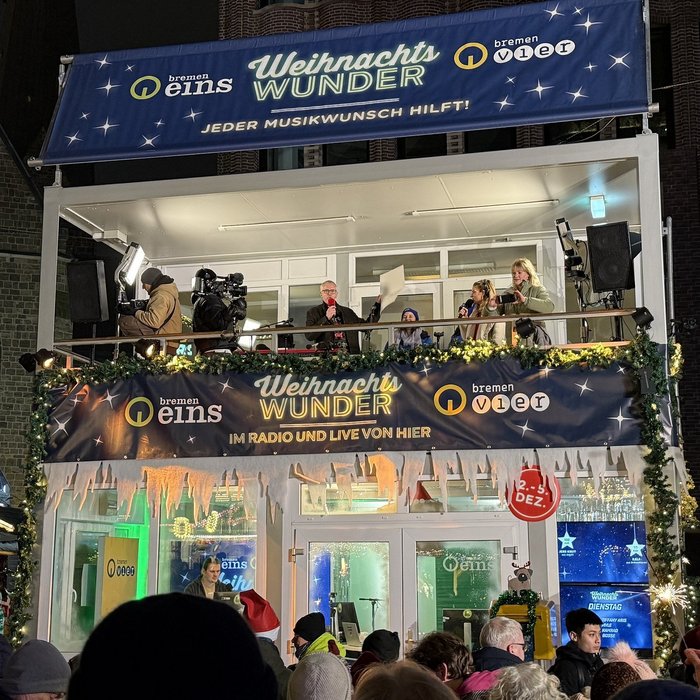 Bild vom Bremer Weihnachtsmarkt. In der Mitte ist das gläserne Studio zu sehen. Die Moderator*innen stehen auf dem Balkon des Studios. Unten stehen viele Menschen. Es ist dunkel und alles ist weihnachtlich geschmückt. 