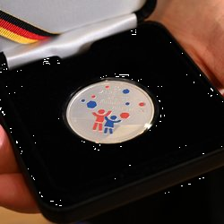 Die Münze zu unserem Jubiläum zeigt unsere Logofiguren sowie rote und blaue sechsecke, die für unsere Spendendose stehen. 